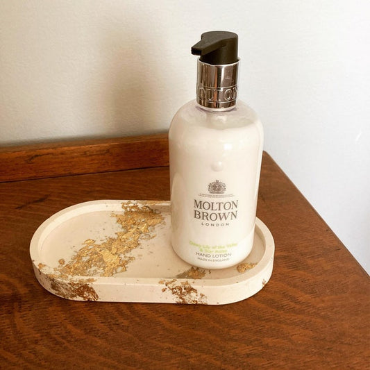 White rectangular tray with rounded edges to make an elongated oval. Gold leaf banding across the body of the tray. Molton Brown hand cream sat on tray which is on top of a dark brown wooden cabinet